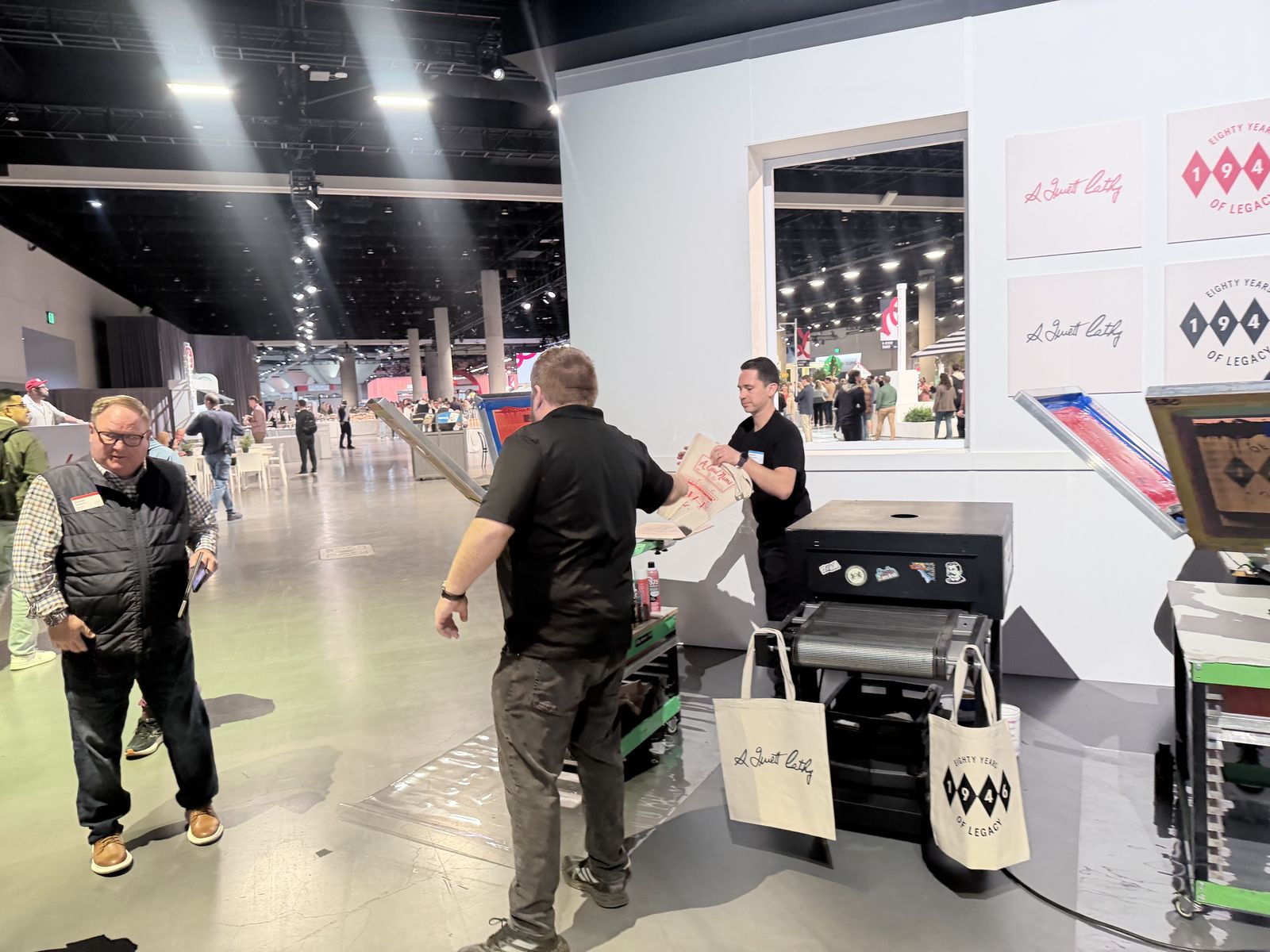 Crew handing off a printed canvas tote next to a conveyor dryer at a convention booth